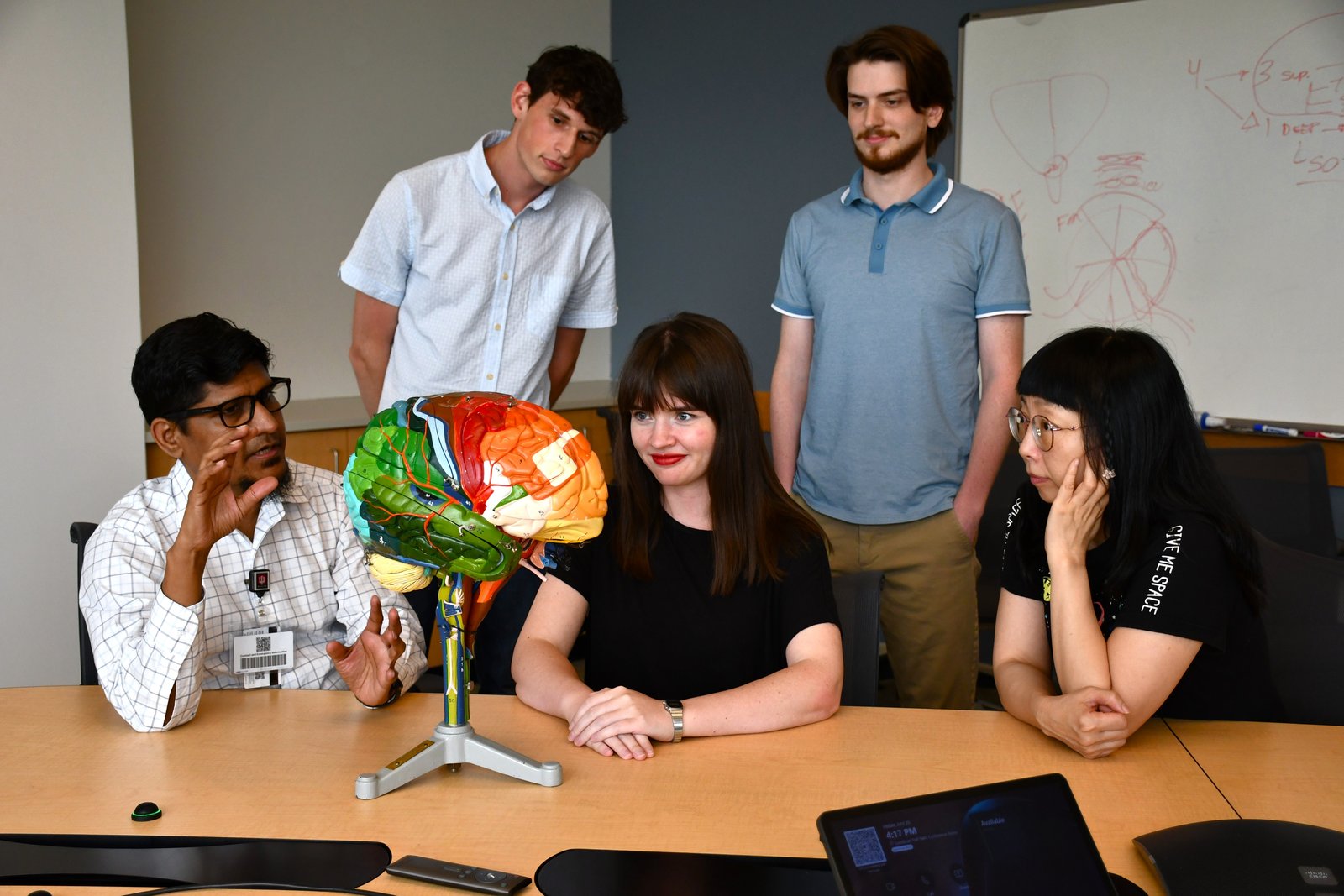 Dr. Noor explaining brain anatomy with a color-coded brain model while four lab members look on around a conference table