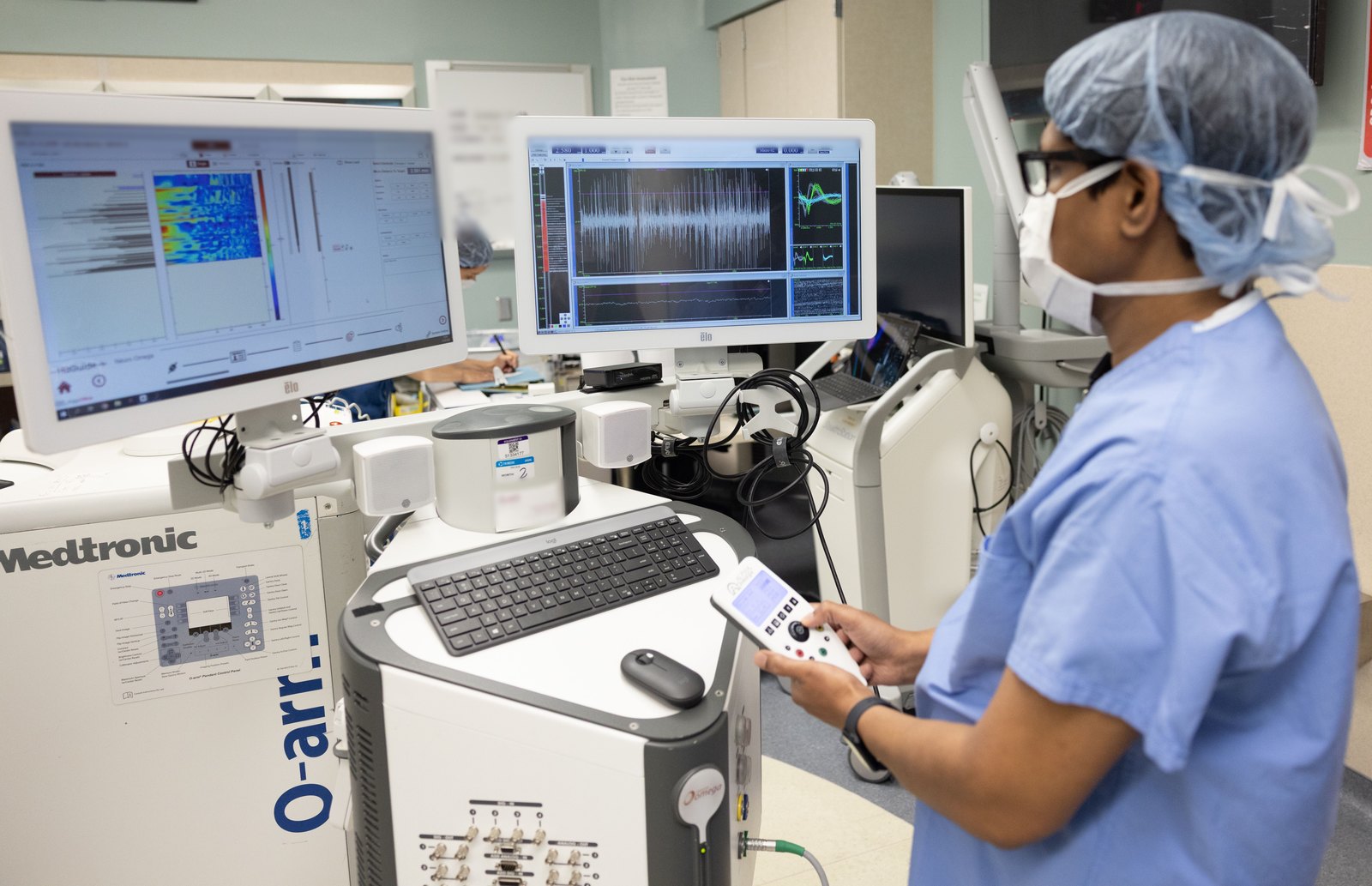 Dr. Noor in surgical scrubs using a handheld controller while monitors display live single-neuron recordings during deep brain stimulation surgery
